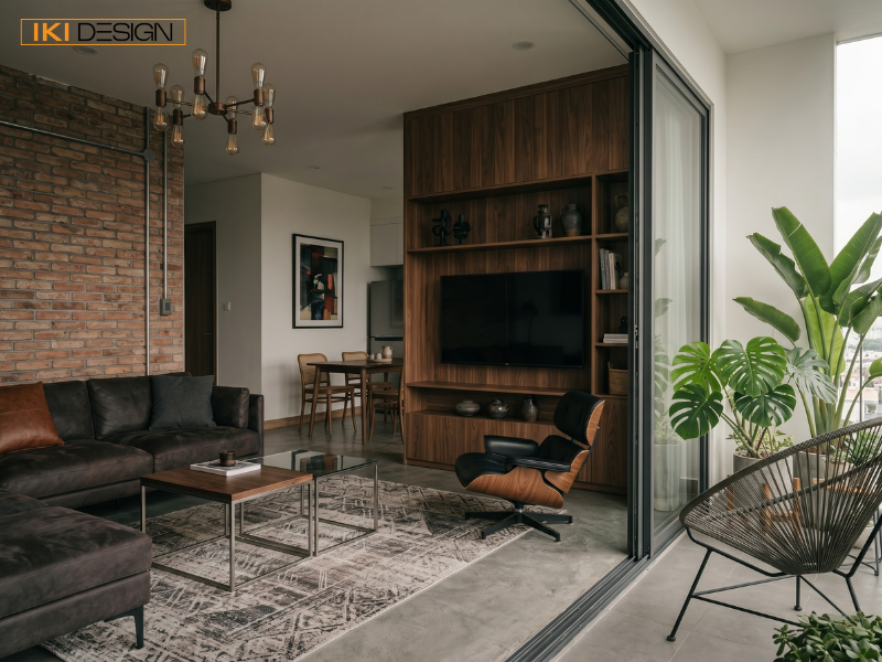 Góc phòng khách với tường gạch trần, sofa da nâu, hệ tủ gỗ lớn, ghế Eames Lounge và ban công ngập tràn cây xanh